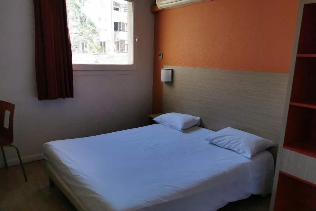 Un lit simple avec une literie blanche, dans une pièce dont un mur est peint en orange, avec une fenêtre dotée de rideaux et une chaise.