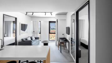 A modern hotel room with a dining area, a sofa, a TV, and a large window offering a city view.