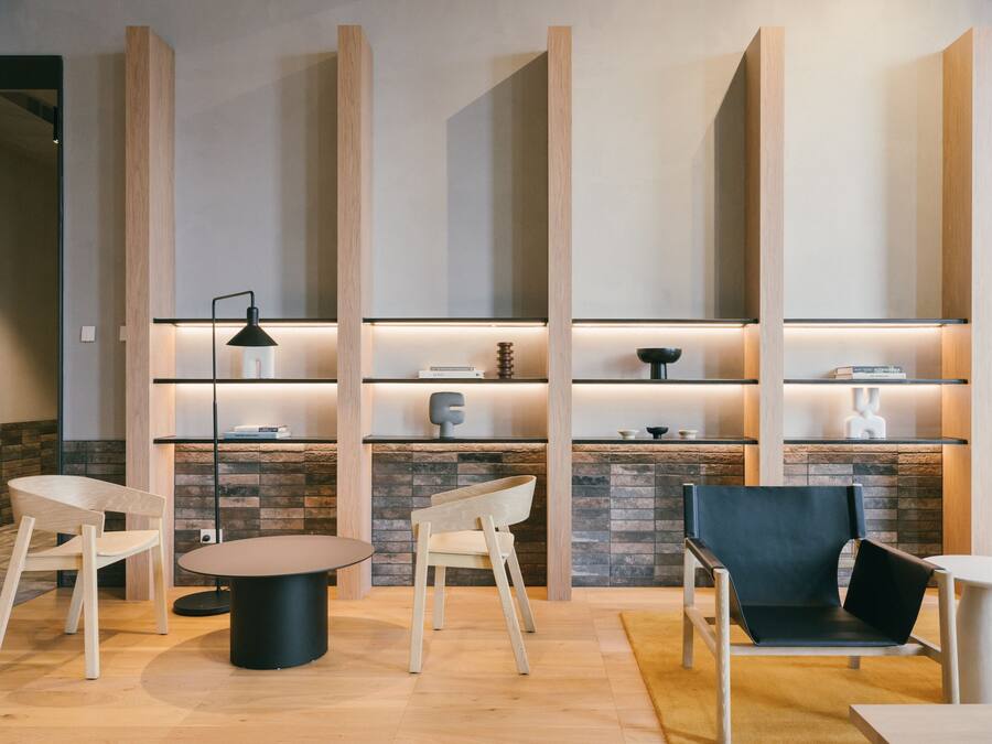 A modern lounge area with wooden chairs, a round table, and a black chair. Shelves with decorative items and a stone wall are in the background.