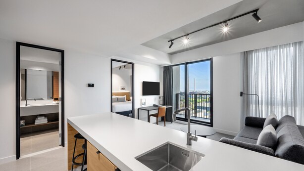 A modern hotel room with a kitchen area, a balcony with a city view, and a large window with curtains. - Nesuto Docklands Apartment Hotel (Docklands)