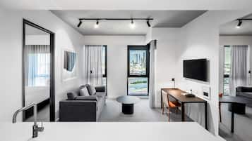 A modern hotel room with a sofa, a coffee table, a TV, and a city view through large windows.