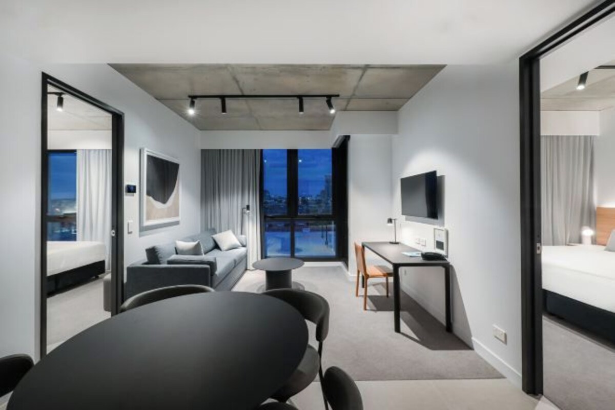 Een moderne hotelkamer met een groot bed, een bureau, een bank en een eettafel. Een groot raam biedt uitzicht op de stadszicht.