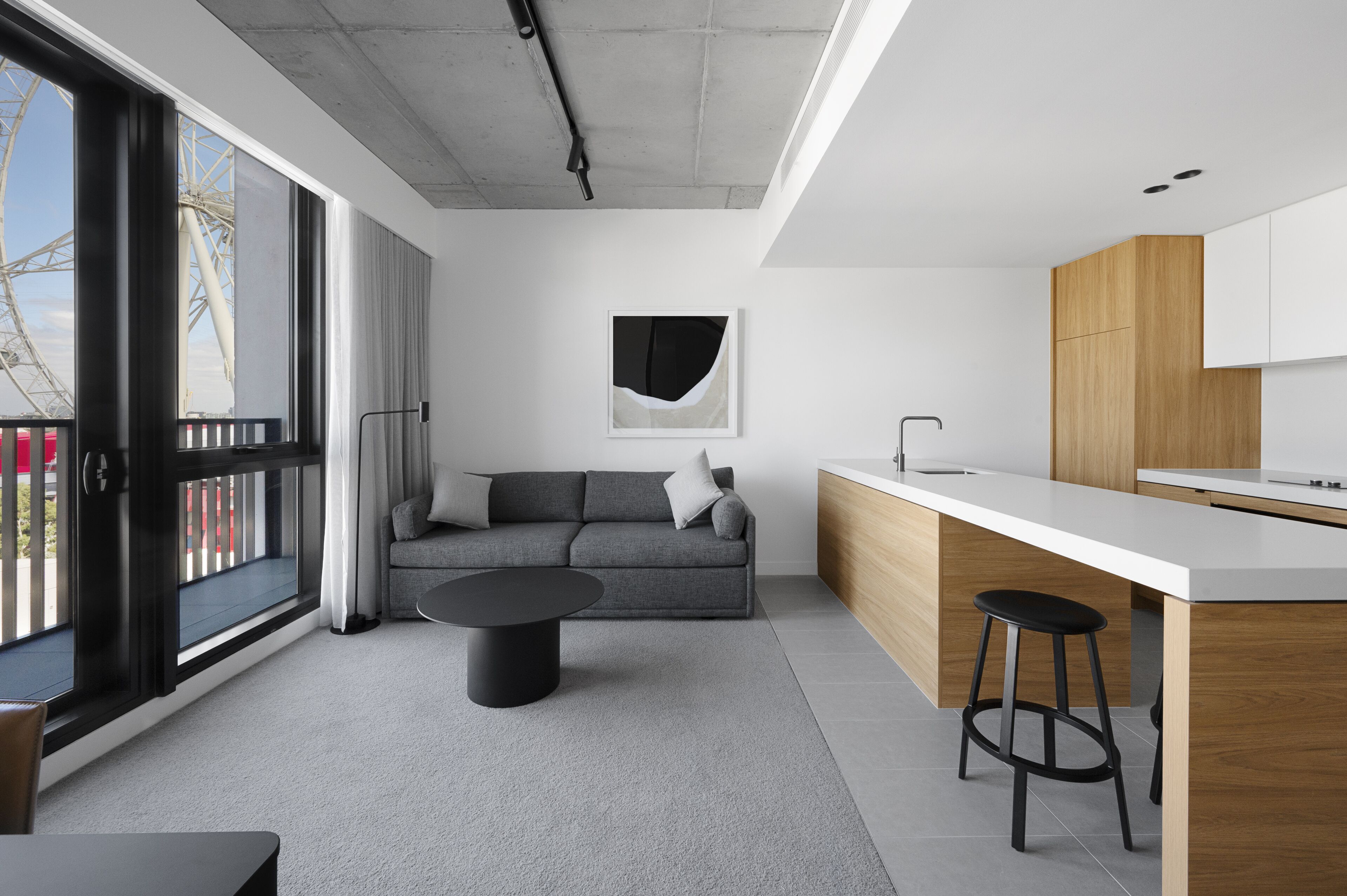 A modern kitchen with a white countertop and wooden cabinets, a gray sofa with a black coffee table, and a large window with a view of a Ferris wheel.