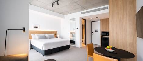 A modern hotel room with a large bed, a dining table with fruit, a sofa, and a small table.