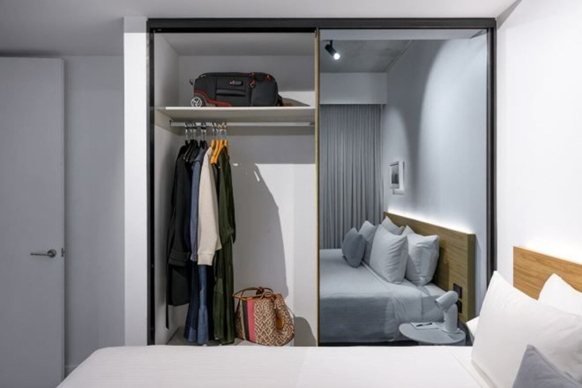 Een hotelkamer met een bed, een kast met kleding eraan, een plank met een tas en een spiegel.