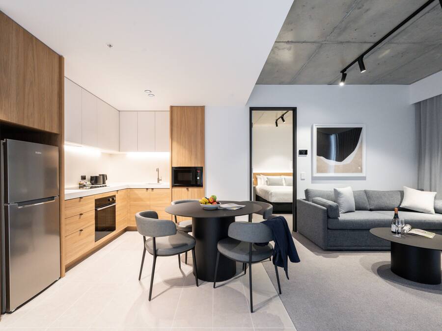 Una cocina moderna con refrigerador, microondas y gabinetes de madera. Hay un comedor con una mesa redonda y sillas, además de un espacio de sala con un sofá y una mesa de centro.