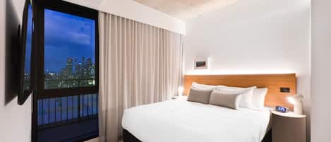 A hotel room with a large bed, a nightstand with a clock, a window with curtains, and a city view.