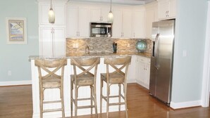 Una cocina con armarios blancos, un refrigerador de acero inoxidable, un protector contra salpicaduras de piedra y tres taburetes de bar de madera.