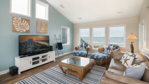 A living room with a large flat-screen TV, wicker furniture, and a view of the ocean through large windows. - Hobbs Realty - Enjoy sunny "Beach Daze" in your private pool and hot tub in this oceanfront home. (Holden Beach)
