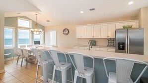 A kitchen with a large island, white bar stools, a stainless steel refrigerator, and a view of the ocean through the windows. - Hobbs Realty - Enjoy sunny "Beach Daze" in your private pool and hot tub in this oceanfront home. (Holden Beach)