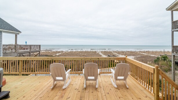 A wooden deck with three white chairs facing a beach. - Hobbs Realty - Enjoy sunny "Beach Daze" in your private pool and hot tub in this oceanfront home. (Holden Beach)
