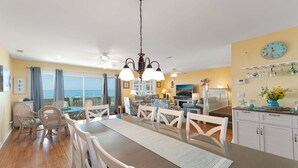 Un comedor con vista al mar, una cocina con un reloj y un balcón con cortinas.
