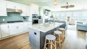 Een moderne keuken met witte kasten, een groot eiland met barstoelen en een zithoek met een grijze bank en tafel.