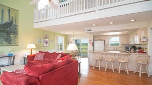 A spacious living area with a red sofa, a dining table, and a kitchen with white cabinets and a bar area. - Hobbs Realty - “Casa Del Mar” | 4BR Marsh-View Home w/ Private Gazebo (Holden Beach)