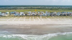 A coastal residential area with beachfront properties, including houses and condos with direct ocean views, a sandy beach, and a grassy field in the background. - Hobbs Realty - “Casa Del Mar” | 4BR Marsh-View Home w/ Private Gazebo (Holden Beach)