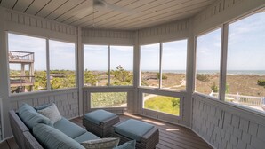 A covered outdoor seating area with wicker furniture and large windows offering a view of the beach and ocean. - Hobbs Realty - Oceanfront with Heated Pool, Elevator, and Hot Tub (Holden Beach)