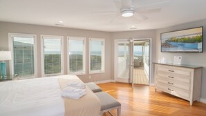 A hotel room with a bed, a bench, a desk, a ceiling fan, and a TV on the wall. - Hobbs Realty - Oceanfront with Heated Pool, Elevator, and Hot Tub (Holden Beach)