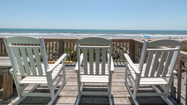 Tres sillas Adirondack blancas en una terraza de madera con vista a una playa y el océano al fondo.