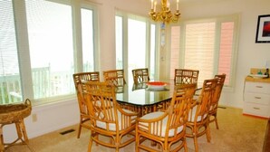 A dining area with a round table surrounded by six wicker chairs, large windows with blinds, and a chandelier. - Hobbs Realty - Enjoy the views in “Clover Reach,” a cute four-bedroom oceanfront home! (Holden Beach)