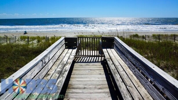 A wooden boardwalk leading to a beach with waves crashing onto the shore. - Hobbs Realty - Enjoy the views in “Clover Reach,” a cute four-bedroom oceanfront home! (Holden Beach)