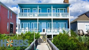 A two-story building with a blue facade and white balconies, surrounded by greenery. - Hobbs Realty - Enjoy the views in “Clover Reach,” a cute four-bedroom oceanfront home! (Holden Beach)