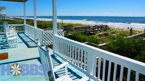 A covered porch with white railings overlooking a beach. - Hobbs Realty - Enjoy the views in “Clover Reach,” a cute four-bedroom oceanfront home! (Holden Beach)