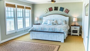A bedroom with a large bed, two bedside tables, a ceiling fan, and a view of the ocean through large windows.