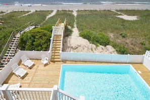 Un área de piscina con muelle de madera, sillas reclinables blancas y vista a la playa.