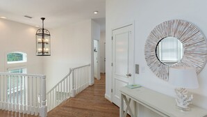A hallway with a staircase, a round mirror, and a table with a lamp. - Hobbs Realty - “Holden 5” | Elegant Stay w/ Elevator, Private Heatable Pool & Panoramic Views (Holden Beach)