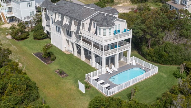 A large, multi-story house with a swimming pool, surrounded by lush greenery and other houses. - Hobbs Realty - “Holden 5” | Elegant Stay w/ Elevator, Private Heatable Pool & Panoramic Views (Holden Beach)