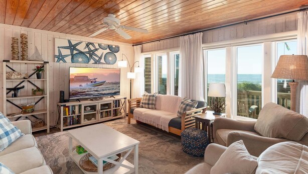 A cozy living room with a wooden ceiling, a television, a sofa, and a view of the ocean.