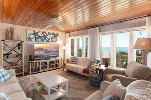 Uma sala de estar aconchegante com teto de madeira, uma televisão, um sofá e vista para o oceano.