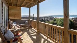 A wooden deck with a railing, overlooking a beach with a wooden pier and a fence.