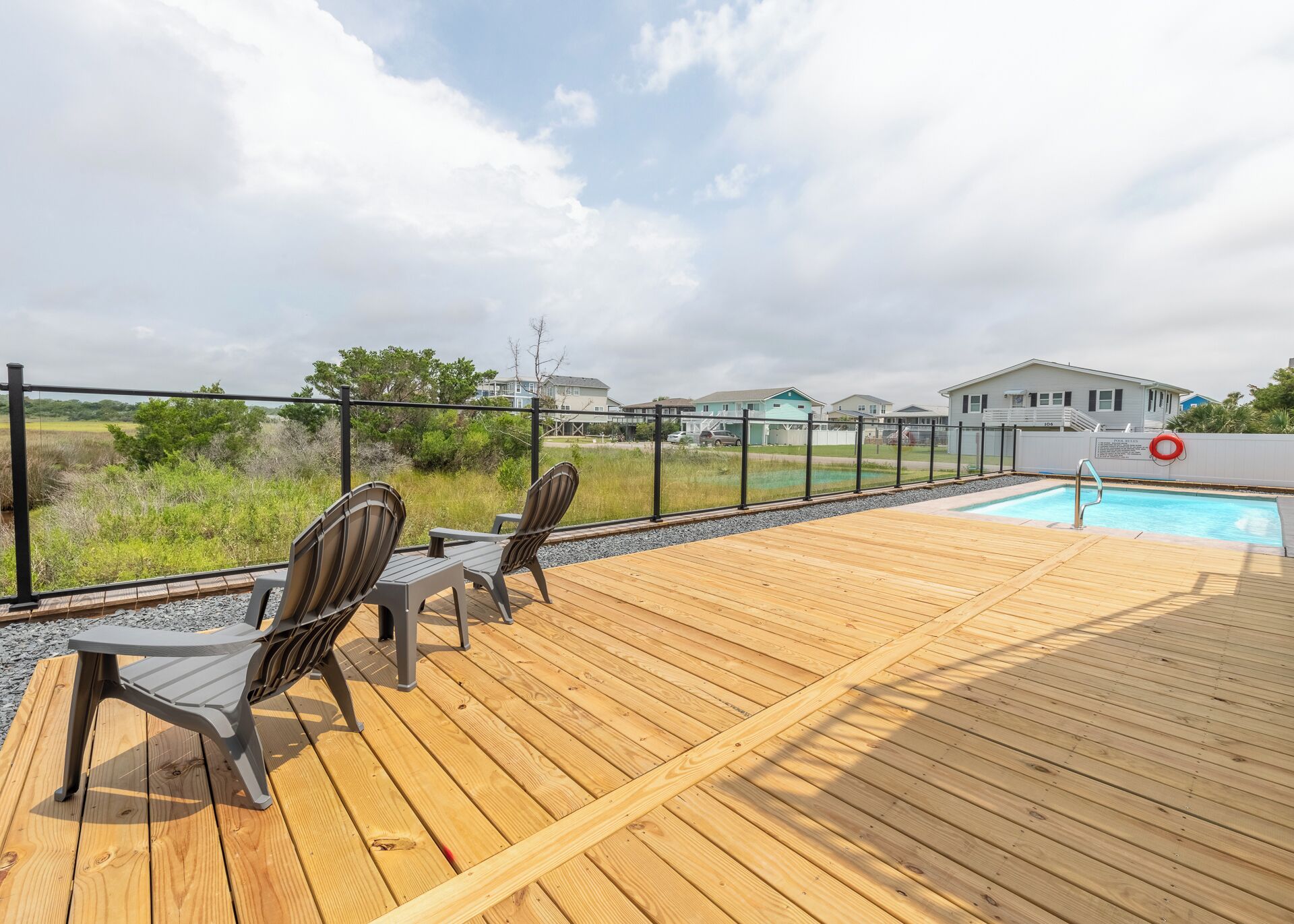 A wooden deck with two Adirondack chairs, a pool, and a view of houses in the background.