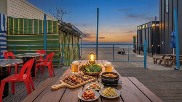 A wooden deck with tables and chairs, a variety of dishes, and a view of the sea at sunset. - Ganghwa Haemolupension (Incheon)