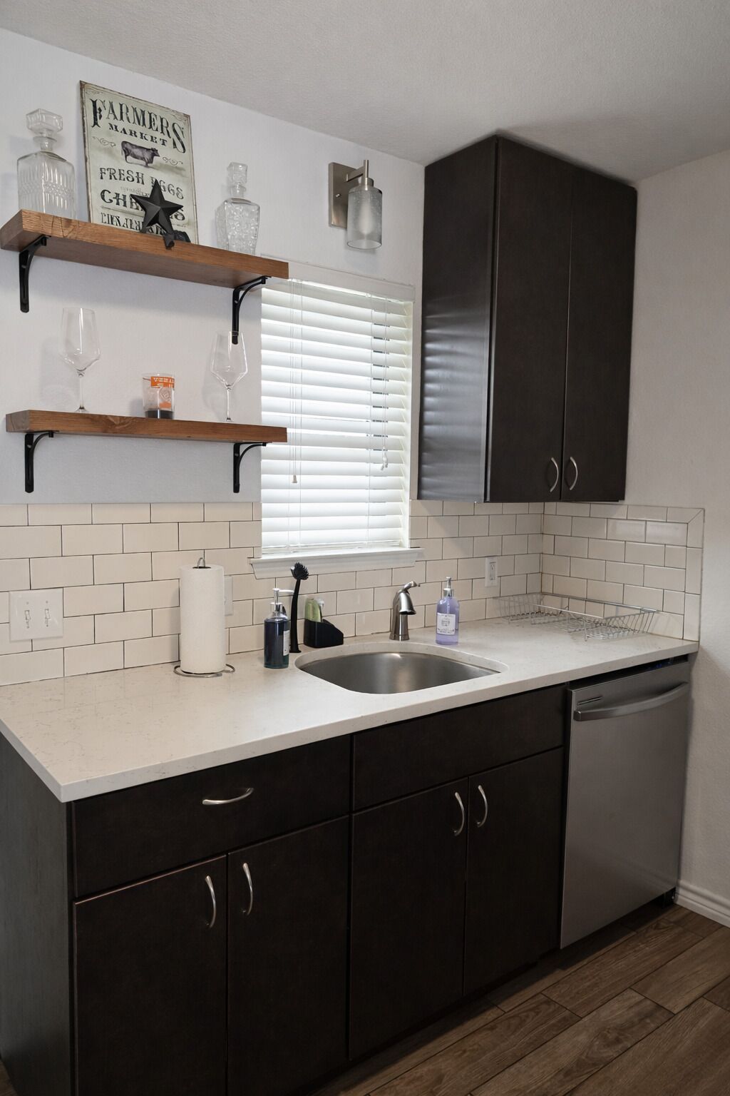Une cuisine avec des meubles de couleur sombre, un plan de travail blanc, un évier en acier inoxydable et une fenêtre avec des stores.