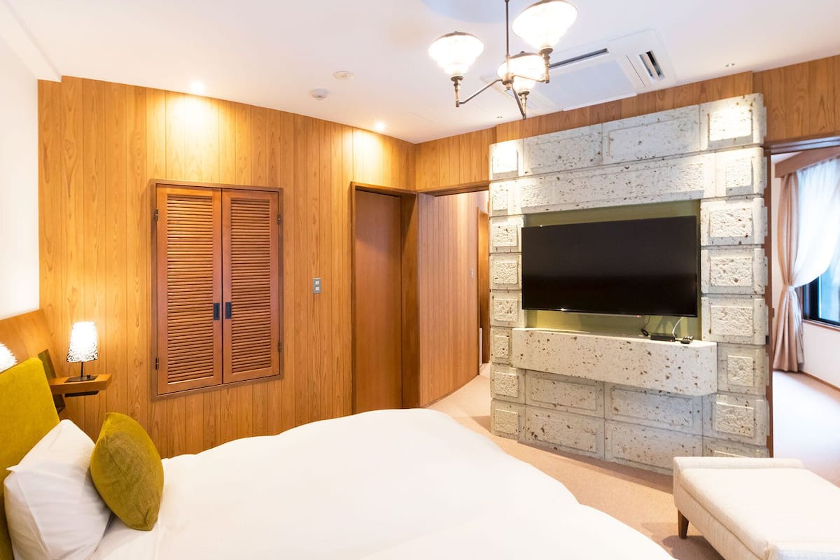Ein Schlafzimmer mit einem Bett, einem an einer Steinwand montierten Fernseher, Holzvertäfelung und einem Fenster mit Vorhängen.