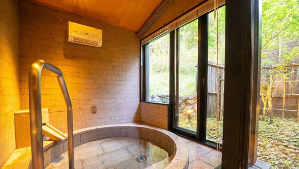 A hot tub with a stone surround, a wooden bench, and a window with a view of greenery. - NIKKO-NISHIMACHICLUB A LA TOUTO (Nikko)