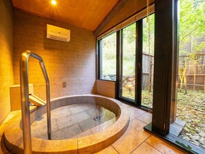 A hot tub with a stone surround, a wooden bench, and a window with a view of greenery.