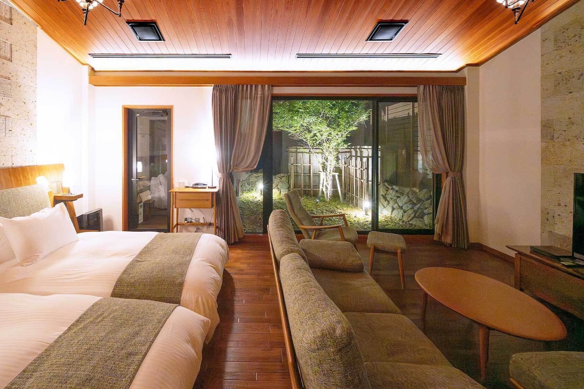 Ein Hotelzimmer mit einem großen Bett, einer Couch, einem kleinen Tisch und einem Fernseher. Eine Glastür führt auf einen Balkon mit Blick auf Grünflächen.