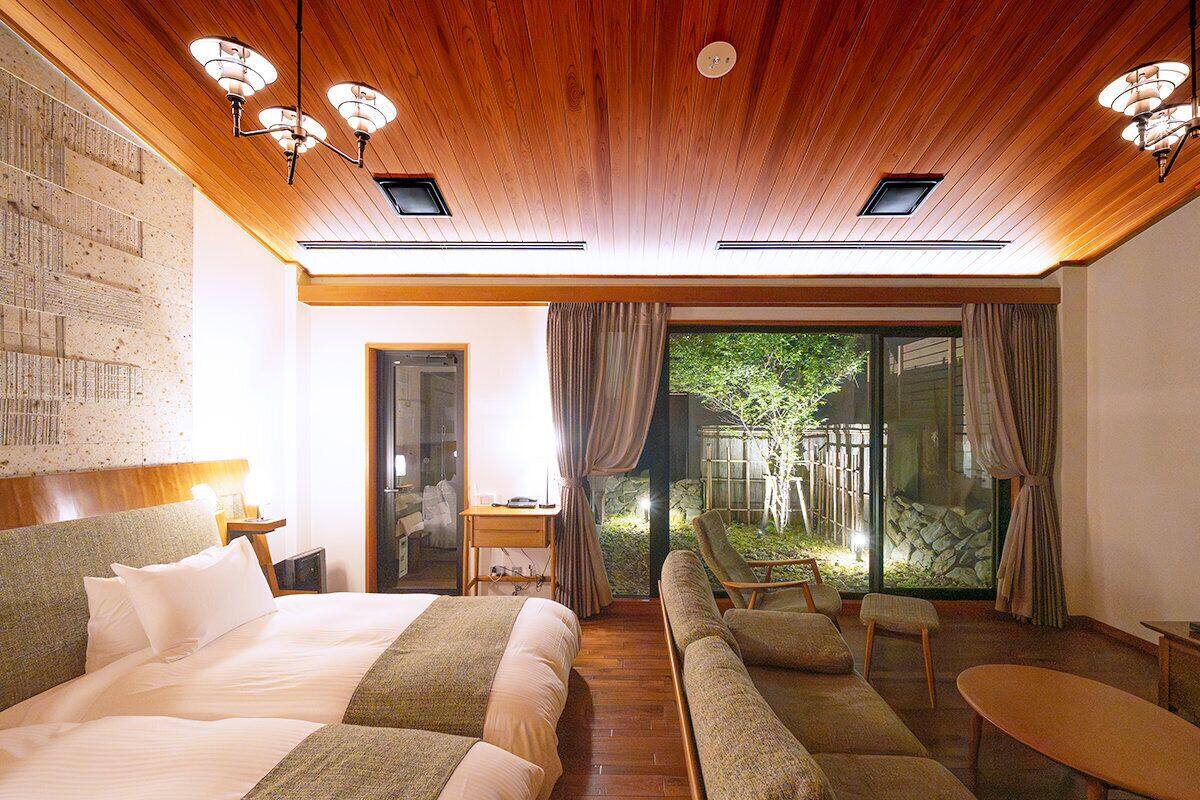 A hotel room with a large bed, a sofa, a small table, and a view of a garden through a glass door.