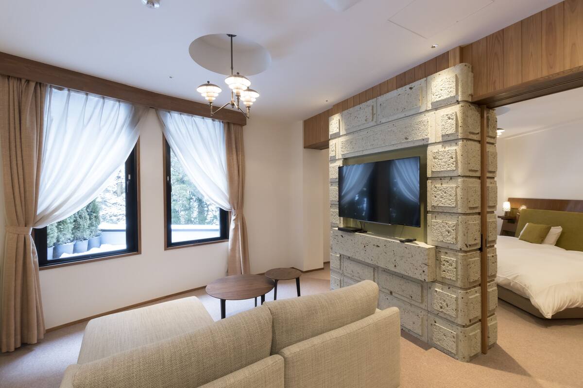 A modern hotel room with a large bed, a seating area with a sofa and two chairs, a TV mounted on a brick wall, and large windows with curtains.