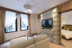 A modern hotel room with a large bed, a seating area with a sofa and two chairs, a TV mounted on a brick wall, and large windows with curtains.