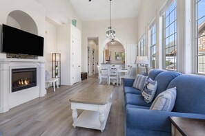 A spacious living room with a fireplace, a large flat-screen TV, a blue sofa, and a dining area with a white table and chairs.