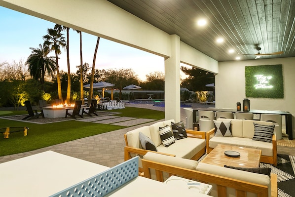 An outdoor patio area with a fire pit, seating, and a pool in the background. - SWSS 7BR THE Place Estate | Pool, Hot Tub, Golf Shed(soon), Volleyball, Putting Green (Scottsdale)
