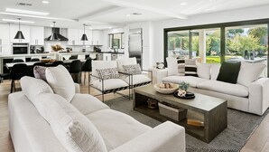 A modern living room with a sofa, armchairs, and a coffee table. There is a kitchen in the background with white cabinets and a dining area. - SWSS 7BR Estate | Golf Sim | Sauna | Pool | Hot Tub | Putting Green | Volleyball | Games (Scottsdale)