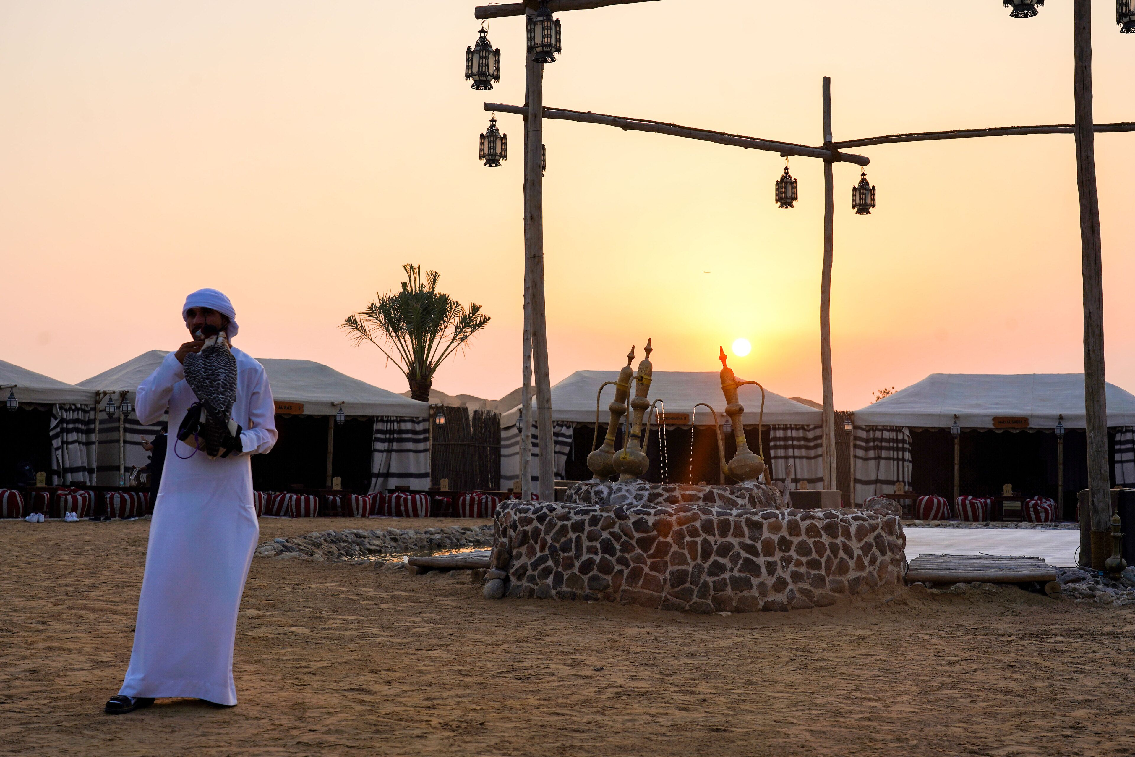 A desert scene with tents and a giraffe enclosure at sunset.