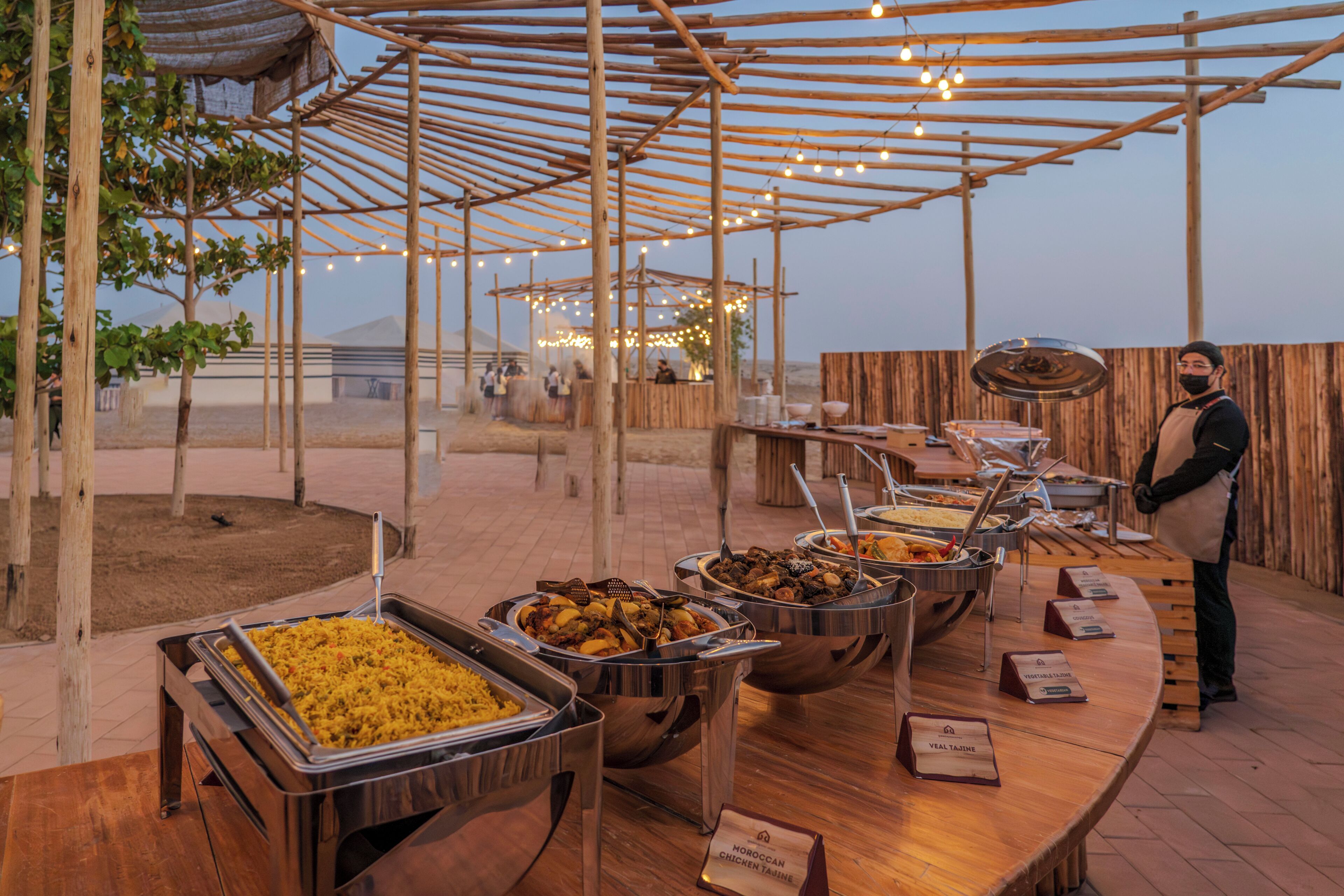 A buffet setup with various food stations, a person standing by the counter, and a wooden pergola structure.