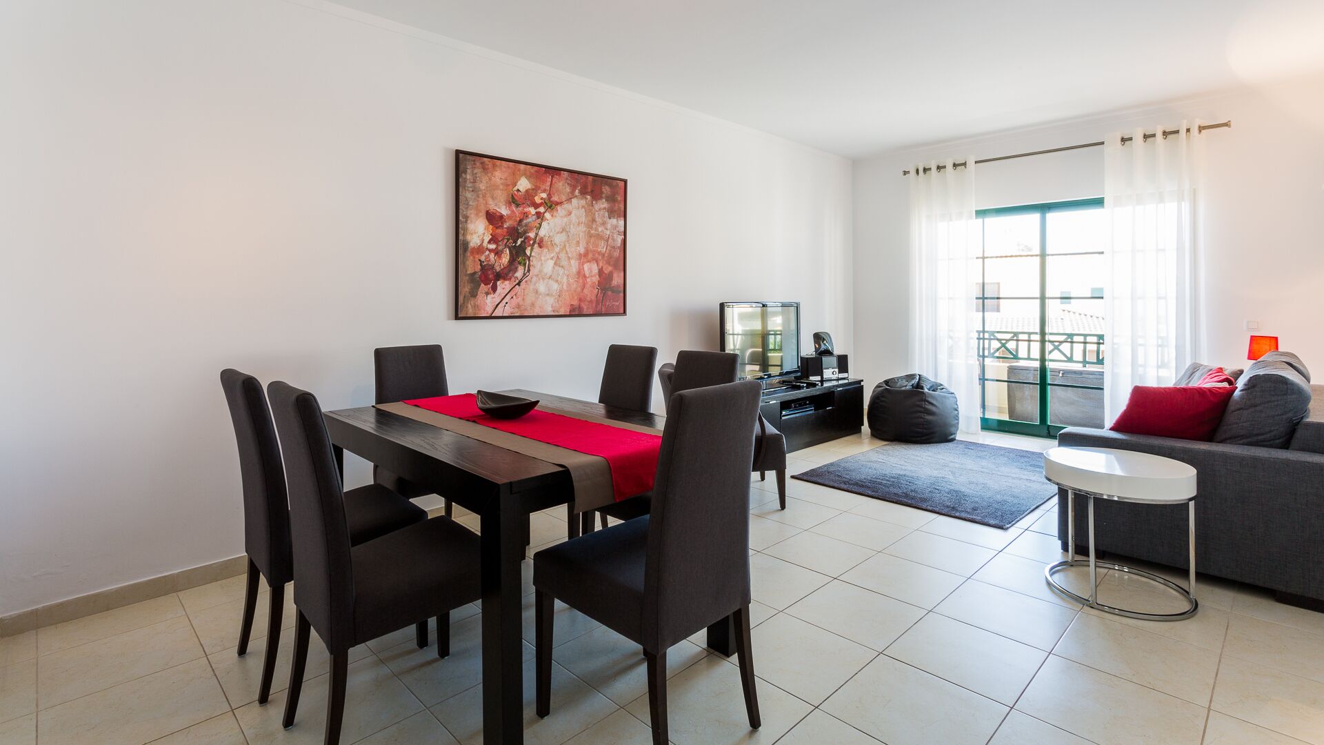 A modern living room with a dining area, a flat-screen TV, and a balcony with a view.