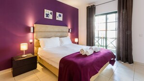 A bedroom with a bed, two wall-mounted lamps, a nightstand, a purple bedspread, and two framed floral artworks. - Central Albufeira 2-B Apt, Sleeps 6, Pool, Parking (Albufeira)